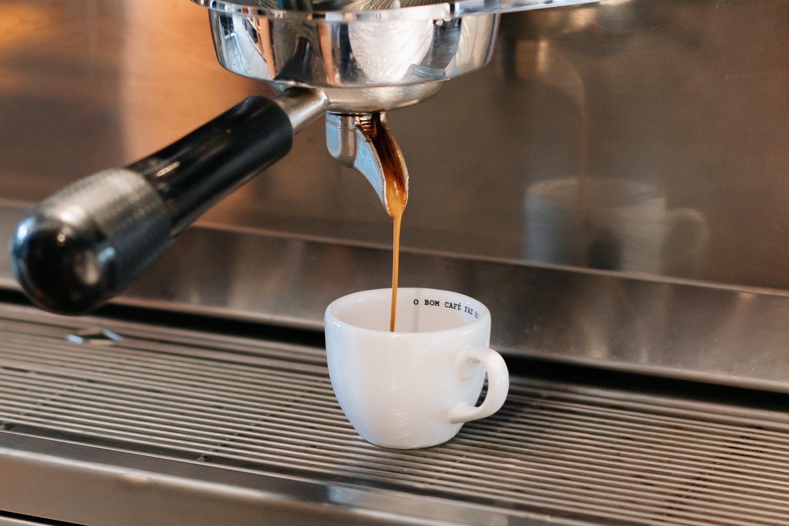 Máquina de café espresso soltando o café em uma xícara branca. Dentro da xícara, é possível ler a mensagem O BOM CAFÉ FAZ BEM.