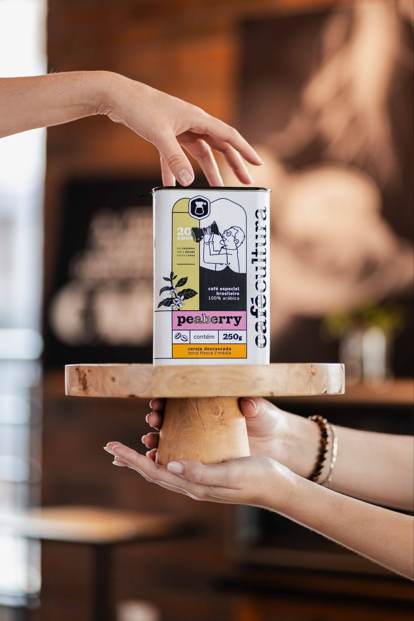 As mãos de duas pessoas seguram um suporte de madeira onde está a lata do blend especial de 20 anos do Café Cultura. 